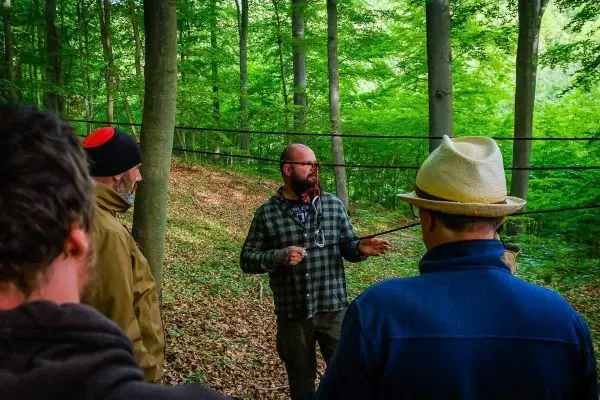 Mann erklärt eine selbstgebaute Seilbrücke im Wald, Teilnehmer hören aufmerksam zu